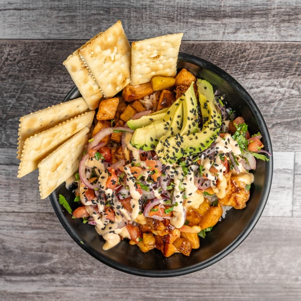 Overhead view of a colorful poke bowl with avocado, red onion, and crackers on a wooden table.
