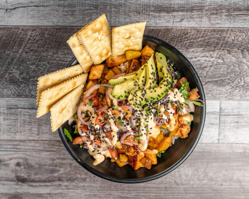 Overhead view of a colorful poke bowl with avocado, red onion, and crackers on a wooden table.