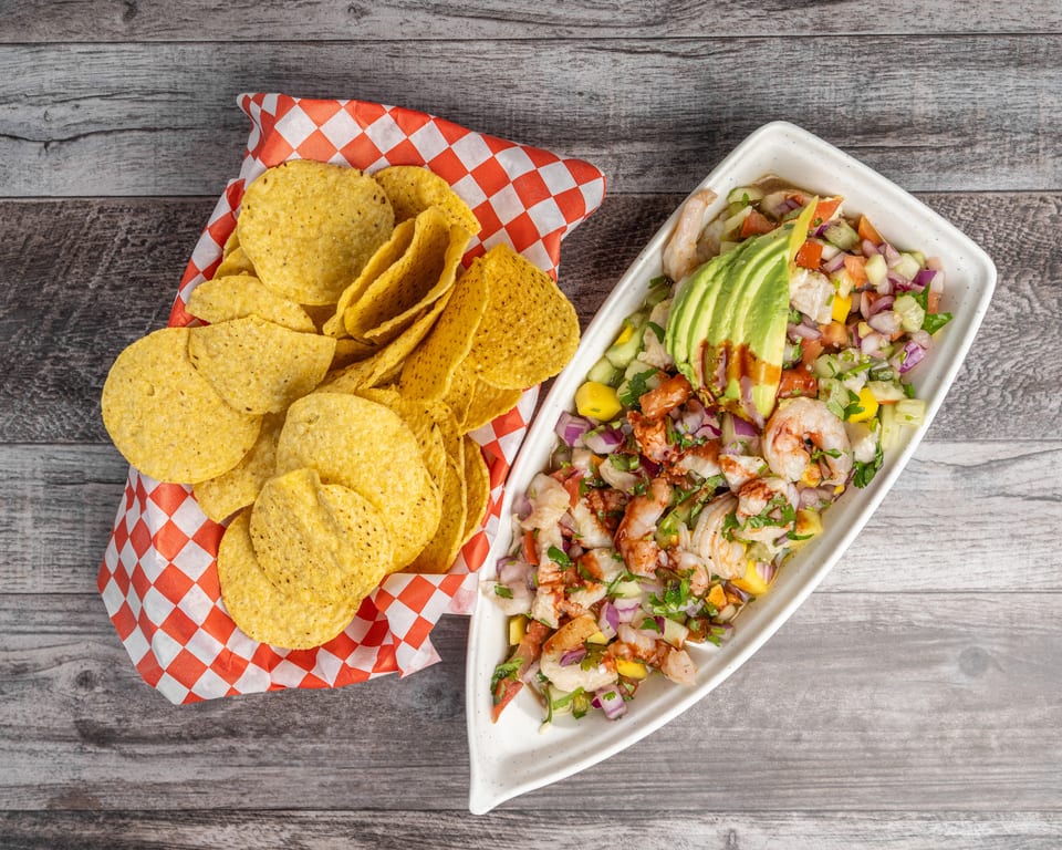 Shrimp ceviche with avocado and salsa in a white dish, next to a basket of tortilla chips.
