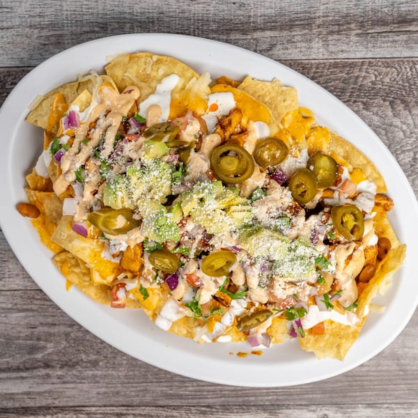 A white oval plate of loaded nachos with cheese, avocado, jalapeños, and sauce on a wooden table.
