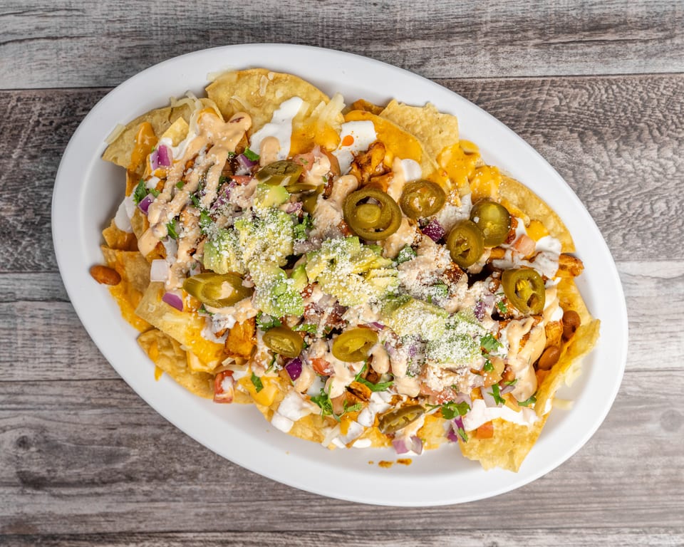 A white oval plate of loaded nachos with cheese, avocado, jalapeños, and sauce on a wooden table.