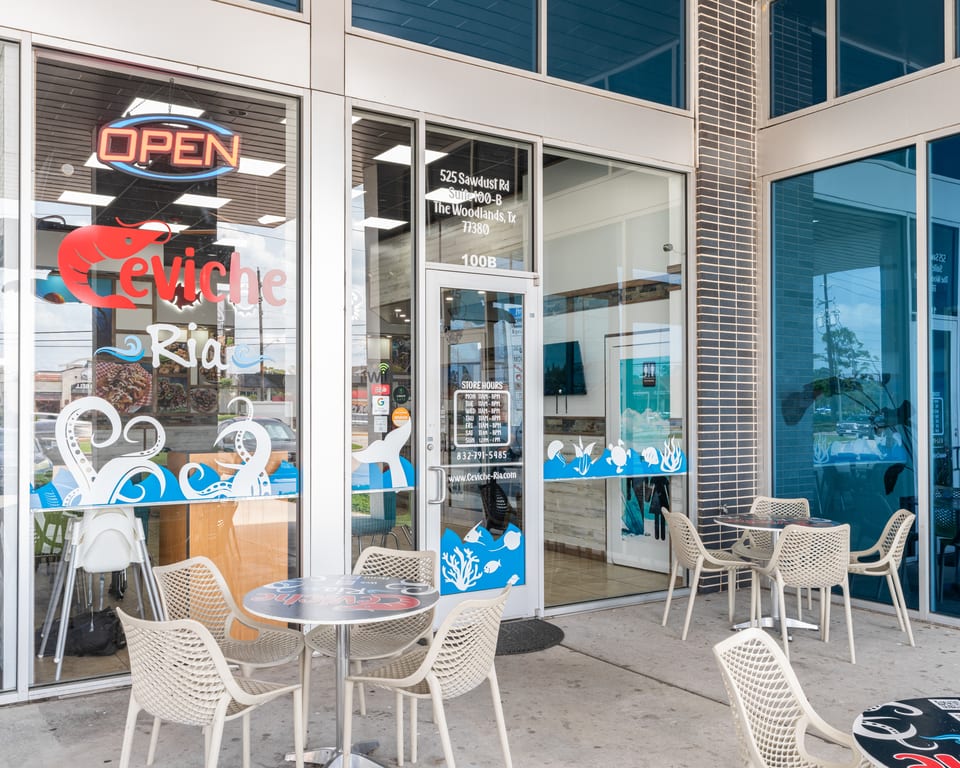 Exterior of Ceviche Ria restaurant with outdoor seating, an "OPEN" sign, and ocean-themed window decals.