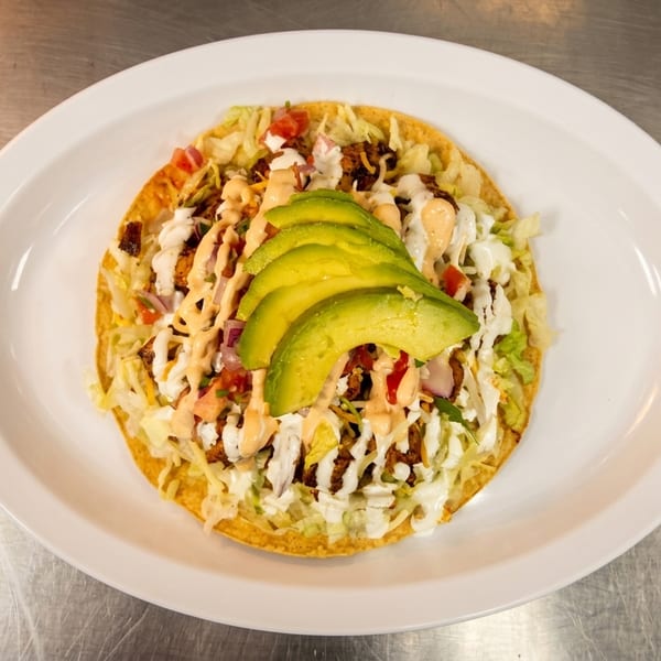 A tostada topped with meat, lettuce, cheese, pico de gallo, sauces, and avocado slices.