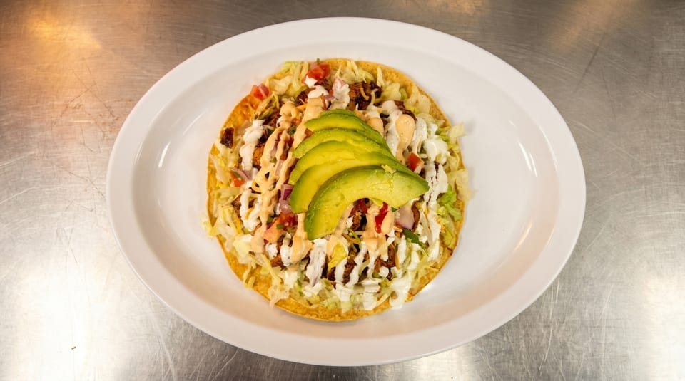 A tostada topped with meat, lettuce, cheese, pico de gallo, sauces, and avocado slices.
