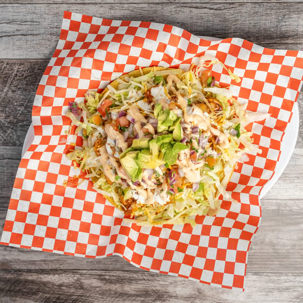 A tostada with shredded lettuce, diced tomatoes, avocado, and sauce on checkered paper.