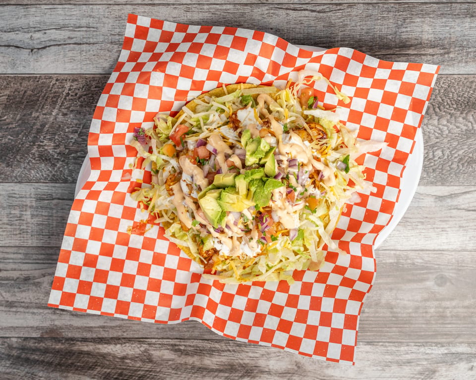 A tostada with shredded lettuce, diced tomatoes, avocado, and sauce on checkered paper.